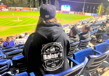 A lady wearing a cap and a bakers drive thru jumper watching a baseball game