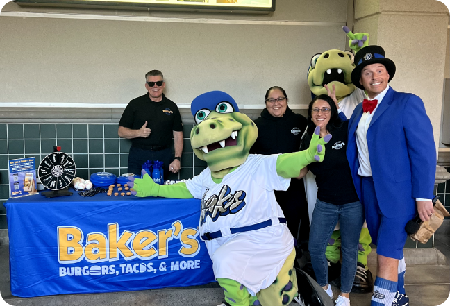 A person in a crocodile suit stands in front of a bakers merchandise table with a man in a blue suit and hat and 3 people from bakers drive thrue