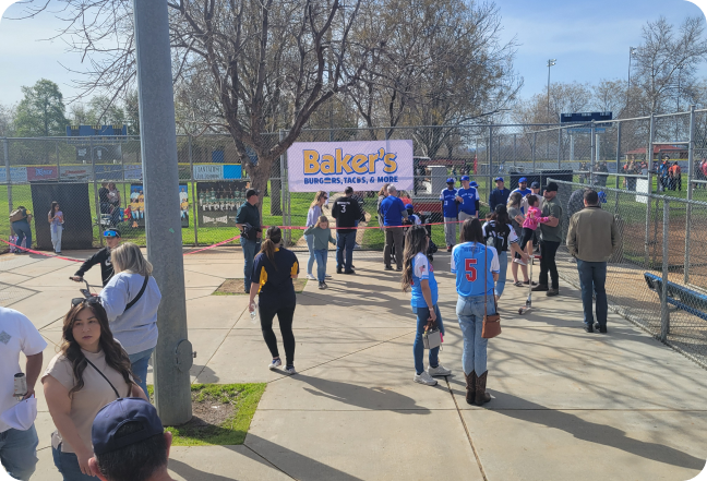 People standing in front of a sign for Bakers drive thru at a baseball game